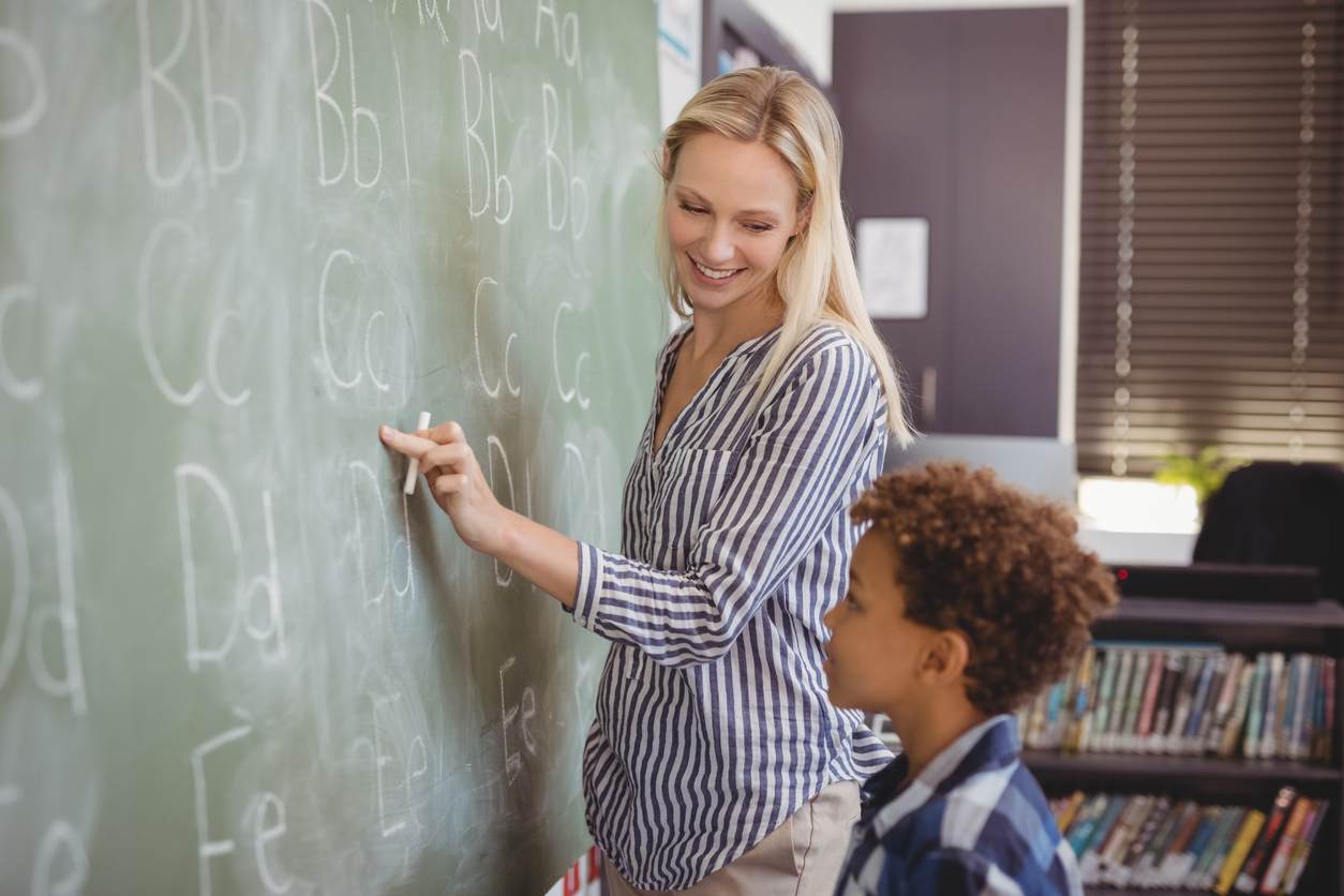 Teacher assisting schoolboy in writing alphabet on chalkboard